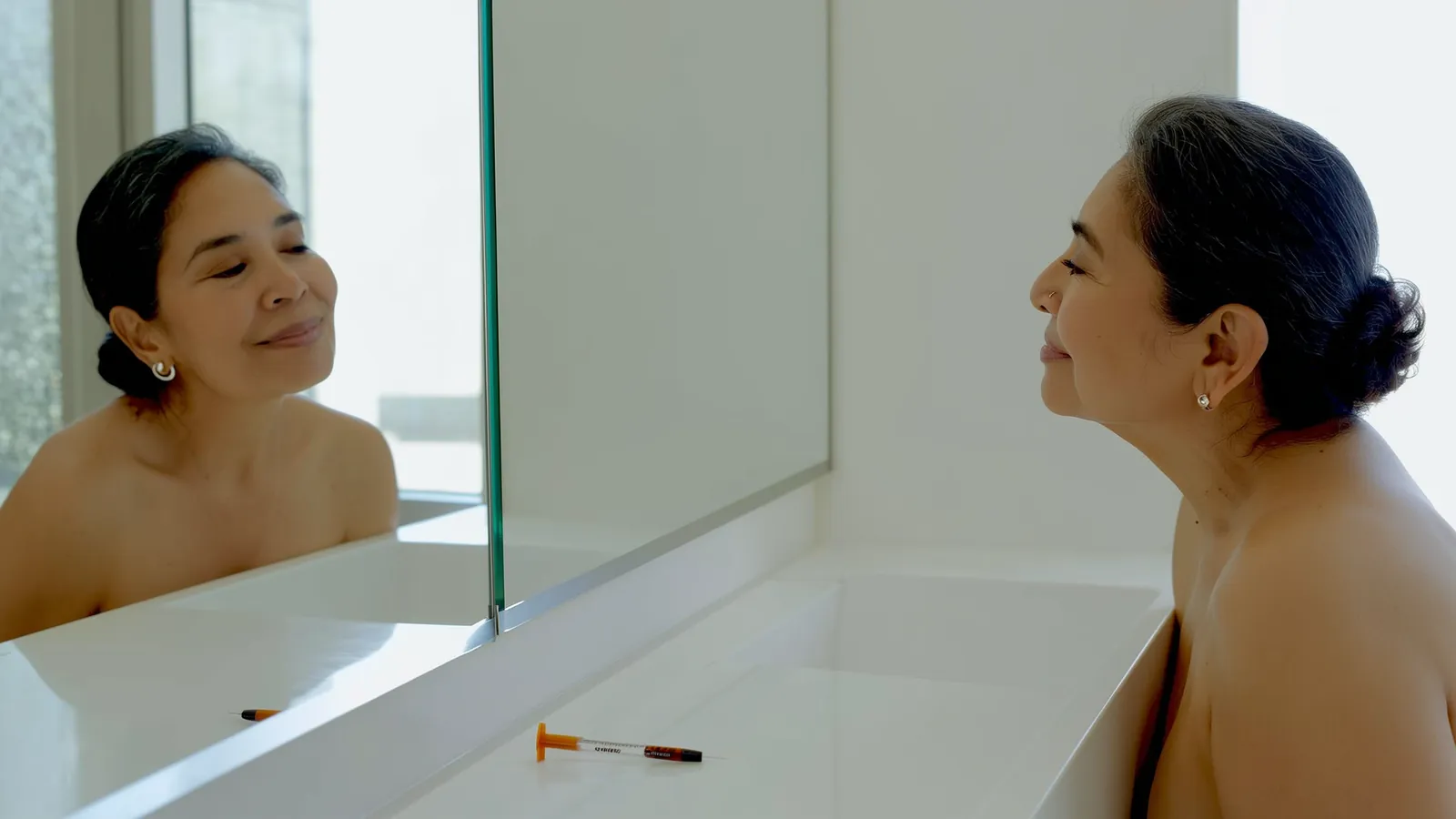 Mujer adulta frente a un espejo observando su rostro con gesto tranquilo, junto a una pluma precargada de Ozempic sobre un tocador, luz natural